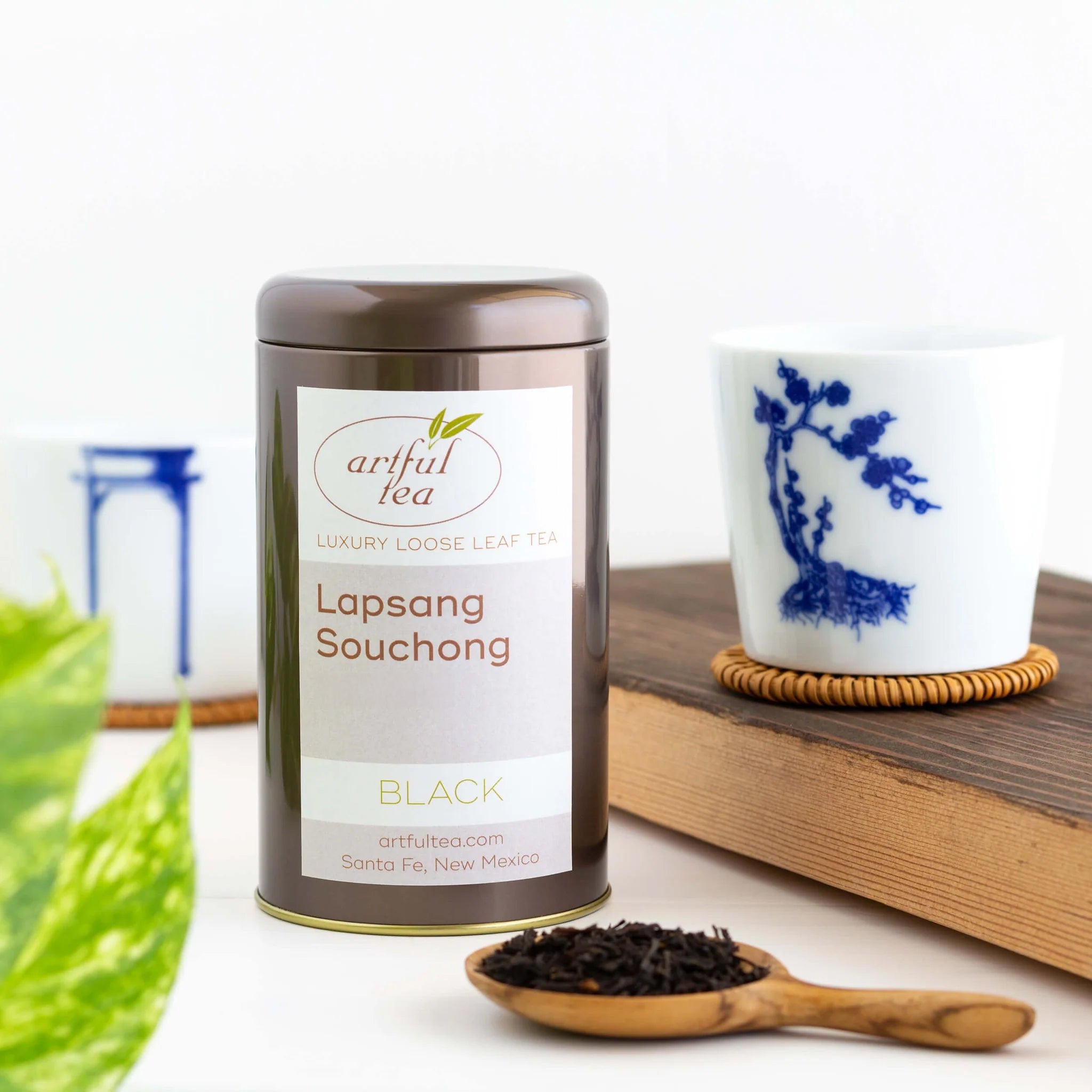 Lapsang Souchong Black Tea shown packaged in a brown tin next to a wooden spoon full of loose leaf tea. Next to it is a wooden plank with a white bonsai cup on top of it on a coaster. In the background is another bonsai cup on top of a coaster.