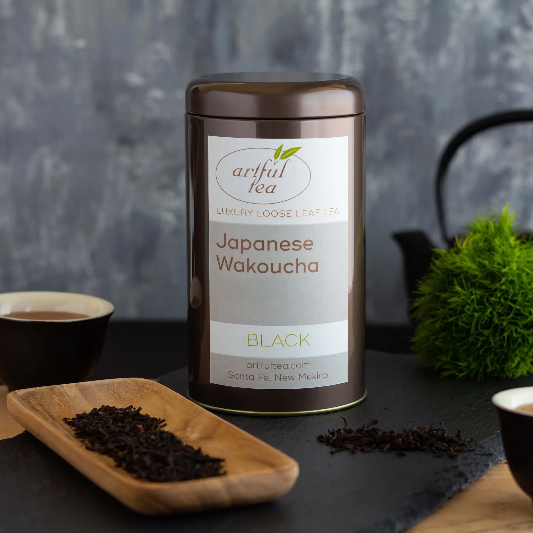 Japanese Wakoucha Black Tea shown packaged in a brown tin alongside a black with white interior full of brewed tea and a wooden platter of loose tea leaves.