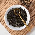 2025 First Flush Darjeeling loose black tea leaves shown in a white bowl with a gold spoon on top of a wooden plank with loose tea leaves beside.