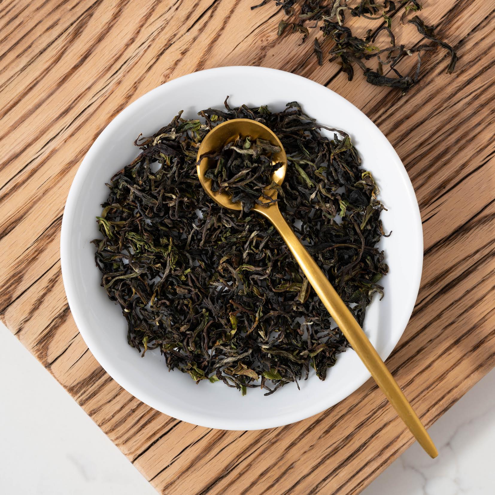 2025 First Flush Darjeeling loose black tea leaves shown in a white bowl with a gold spoon on top of a wooden plank with loose tea leaves beside.