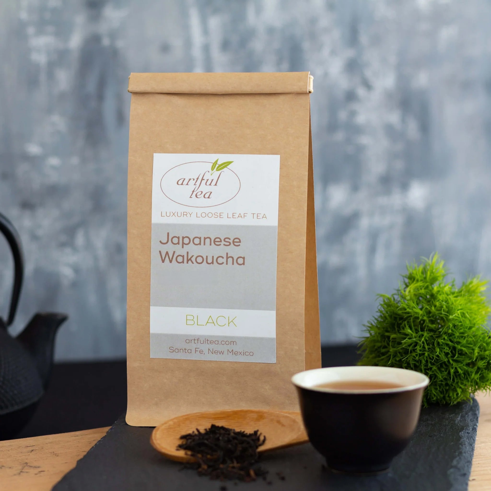 Japanese Wakoucha Black Tea shown packaged in a brown Kraft bag alongside a wooden scoop of loose tea leaves and a black cup full of brewed tea.