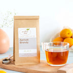 Apricot Brandy Black Tea shown packaged in a brown Kraft bag next to a clear mug of brewed tea and a silver teaspoon of loose leaf tea all on top of a wooden tray with ingredients nearby.