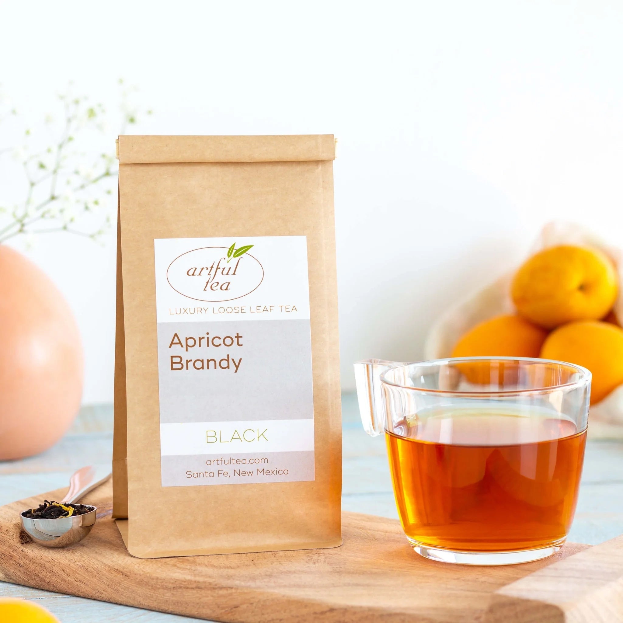 Apricot Brandy Black Tea shown packaged in a brown Kraft bag next to a clear mug of brewed tea and a silver teaspoon of loose leaf tea all on top of a wooden tray with ingredients nearby.